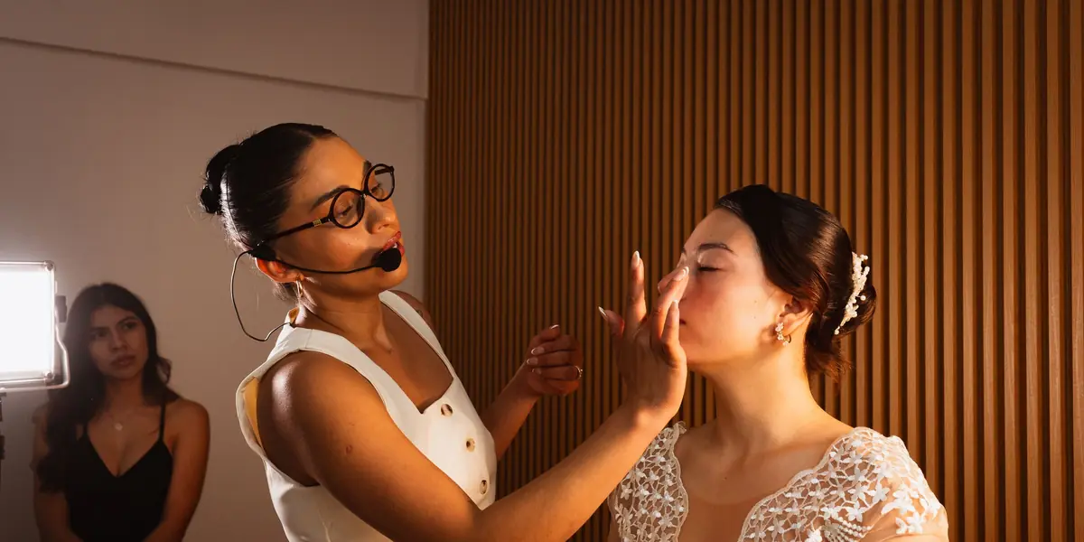 Instructora con anteojos y micrófono de vincha trabajando sobre el rostro de una modelo mientras una alumna mira desde atrás