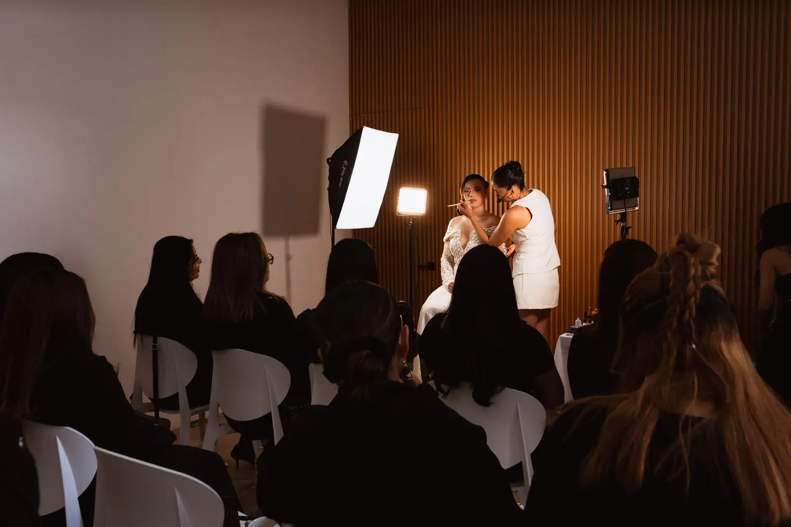Aula llena de alumnas sentadas de espaldas mirando a la instructora y a la modelo al frente, entre luces de estudio