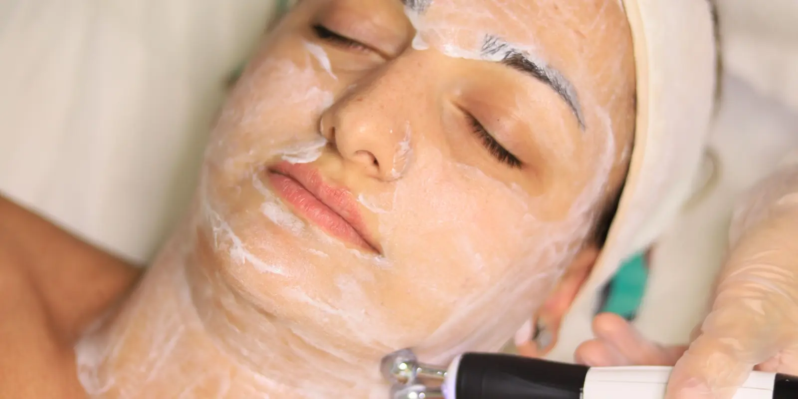 Rostro de una clienta con los ojos cerrados y crema en toda la cara, mientras la cosmetóloga le pasa un cabezal por la mandíbula