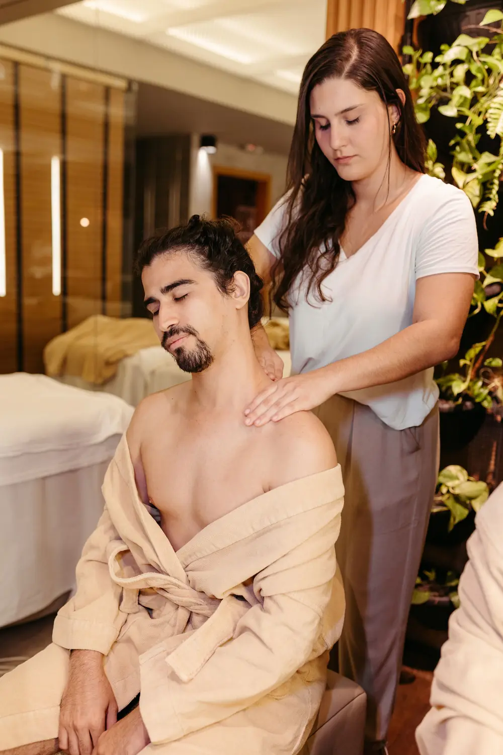 Masajista de pelo castaño masajeando los hombros de un cliente sentado con la bata baja y los ojos cerrados, junto a la camilla