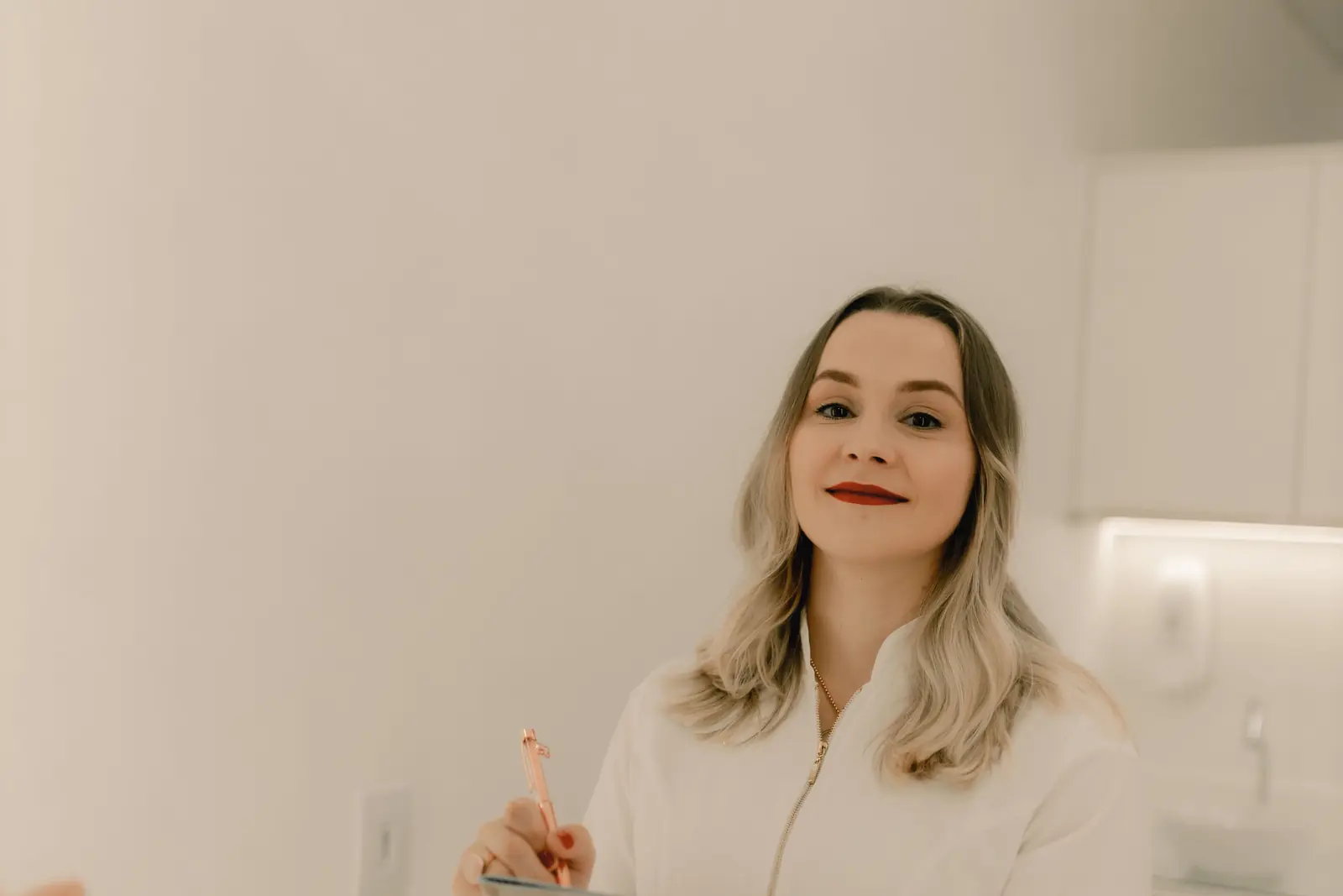 Médica de saco blanco que sonríe a cámara con una tableta en la mano, de pie en su consultorio blanco