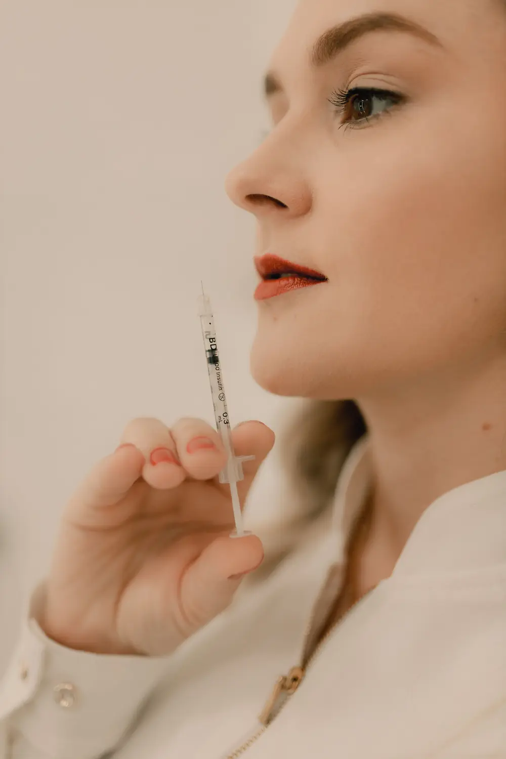Médica de perfil, con el pelo suelto y saco blanco, que sostiene una jeringa descartable a la altura del mentón