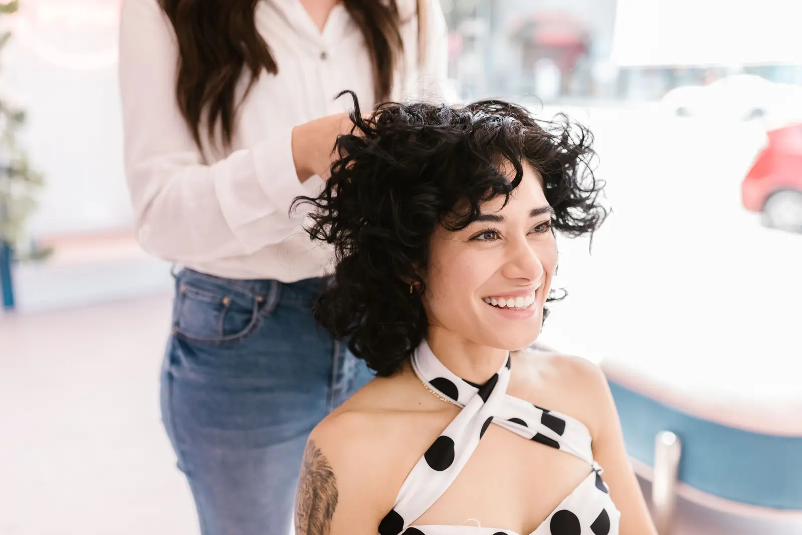 Clienta de pelo negro con rulos sonriendo en el sillón del salón mientras la peluquera, de camisa blanca, le acomoda el pelo